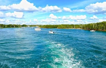 Several motorboats travelling on a turquoise waterway lined with green trees under a partly cloudy sky.
