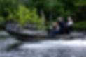 People riding in a small boat on a calm river lined with green trees.