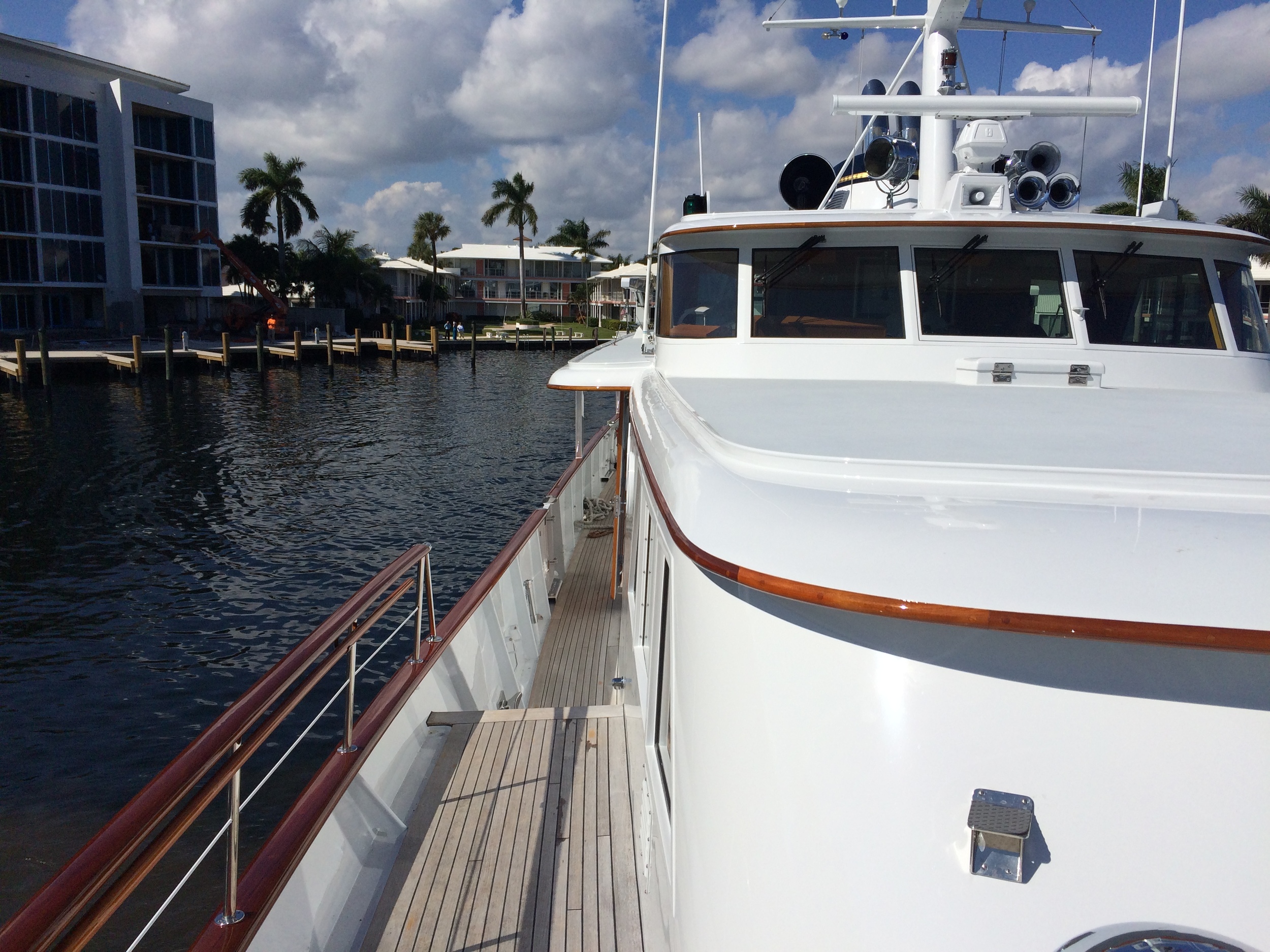 Teak side deck and wood-trimmed rails of a white motor yacht moored along a palm-lined canal with waterfront condominiums beyond.