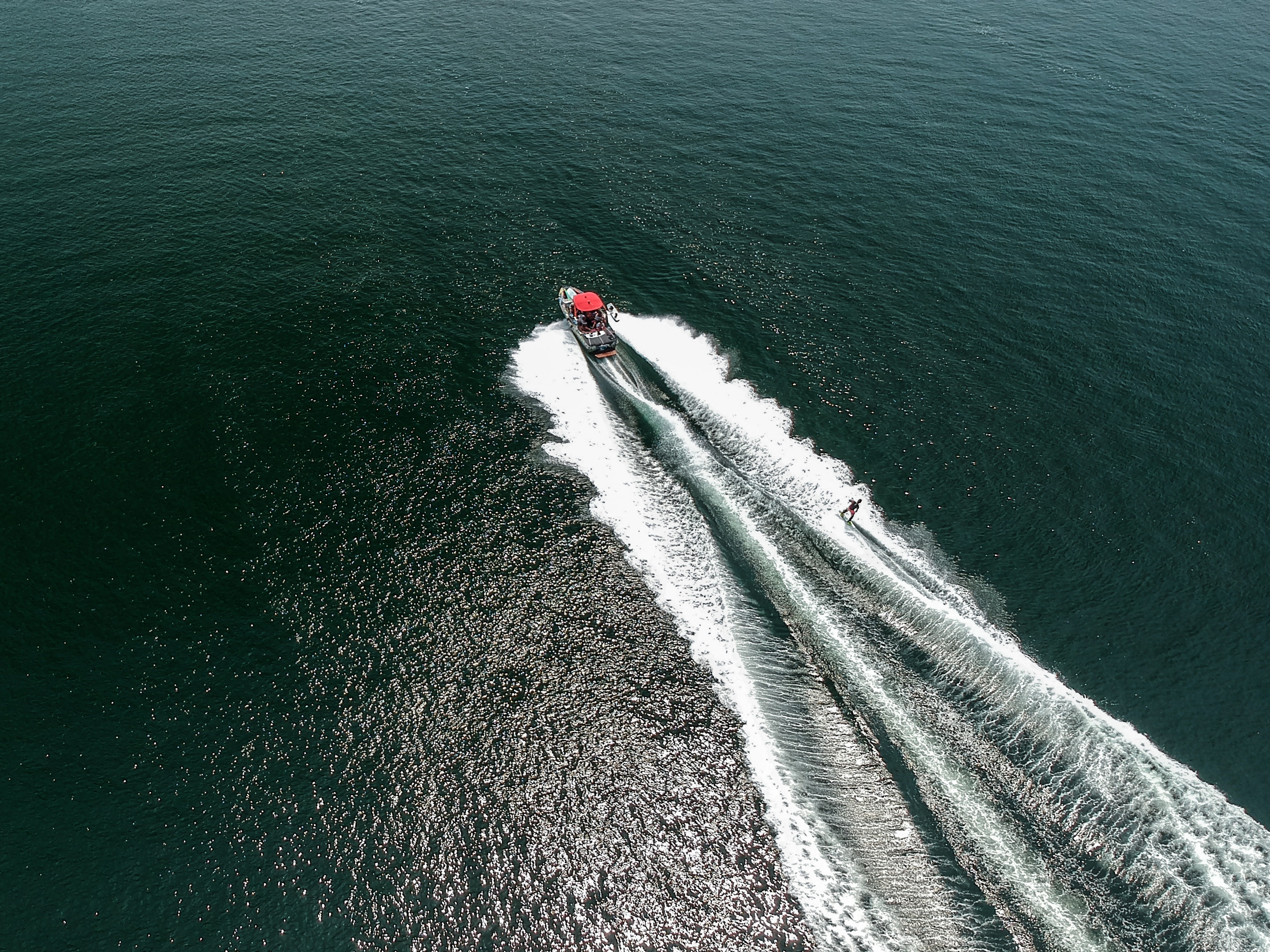 Aerial view of a red-canopy motorboat towing a water skier across open water, leaving white wakes.