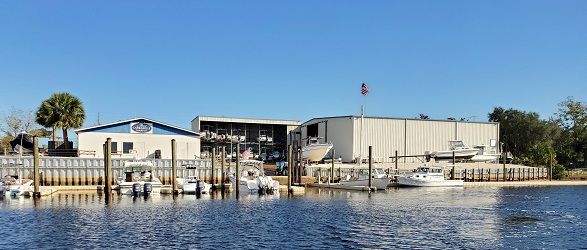 Waterfront marina with docked powerboats and dry-storage buildings along a river, with a flag flying over the property.