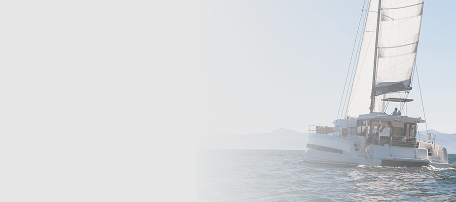 White sailing catamaran with its mainsail raised cruising across open water toward distant mountains.