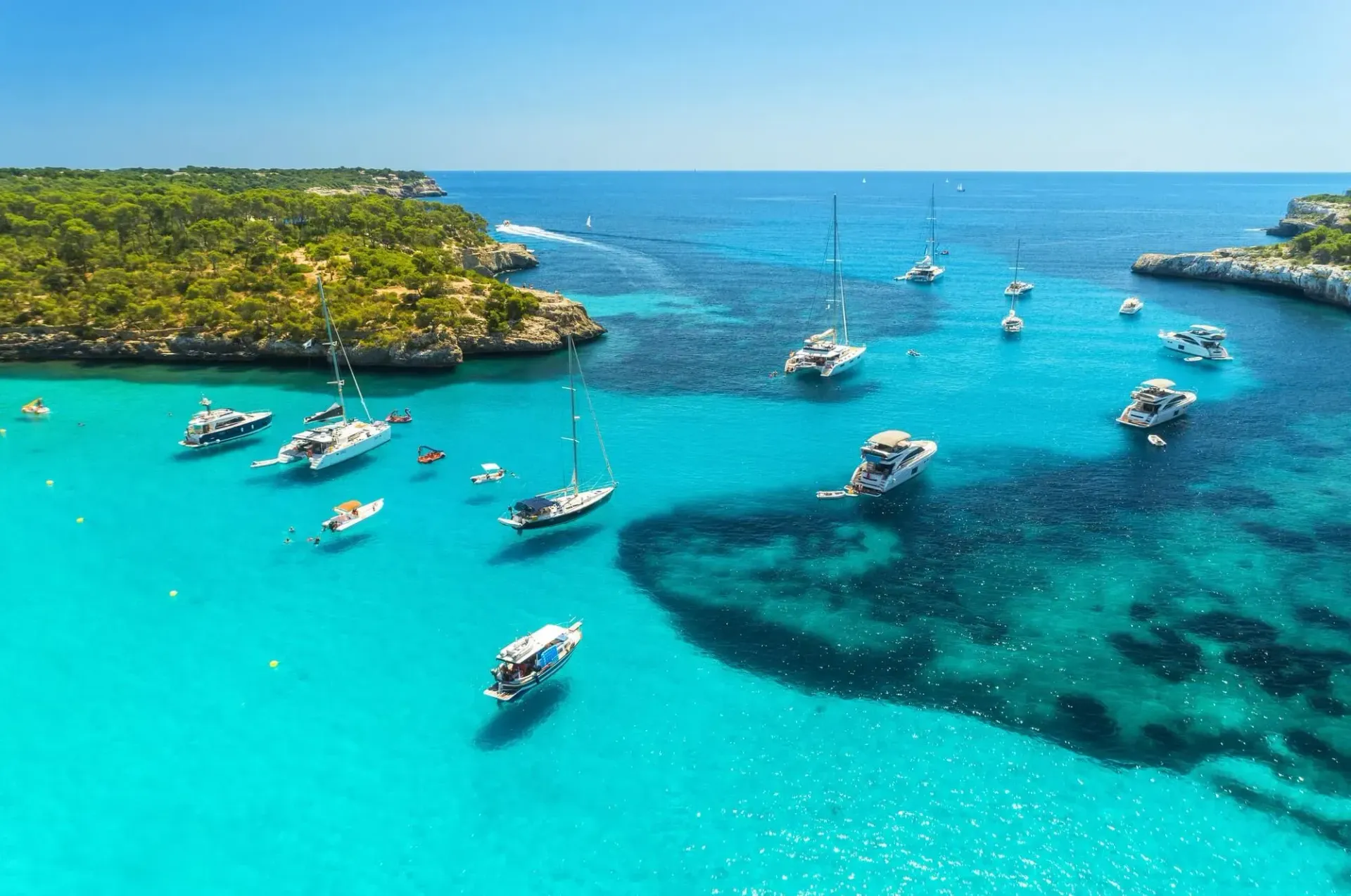 Aerial view of anchored yachts and sailboats in a turquoise cove fringed by pine-covered cliffs, likely Cala Mondragó, Mallorca.