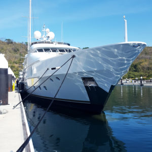 Large white motor yacht moored alongside a concrete dock with mooring lines secured, green hillside behind it.