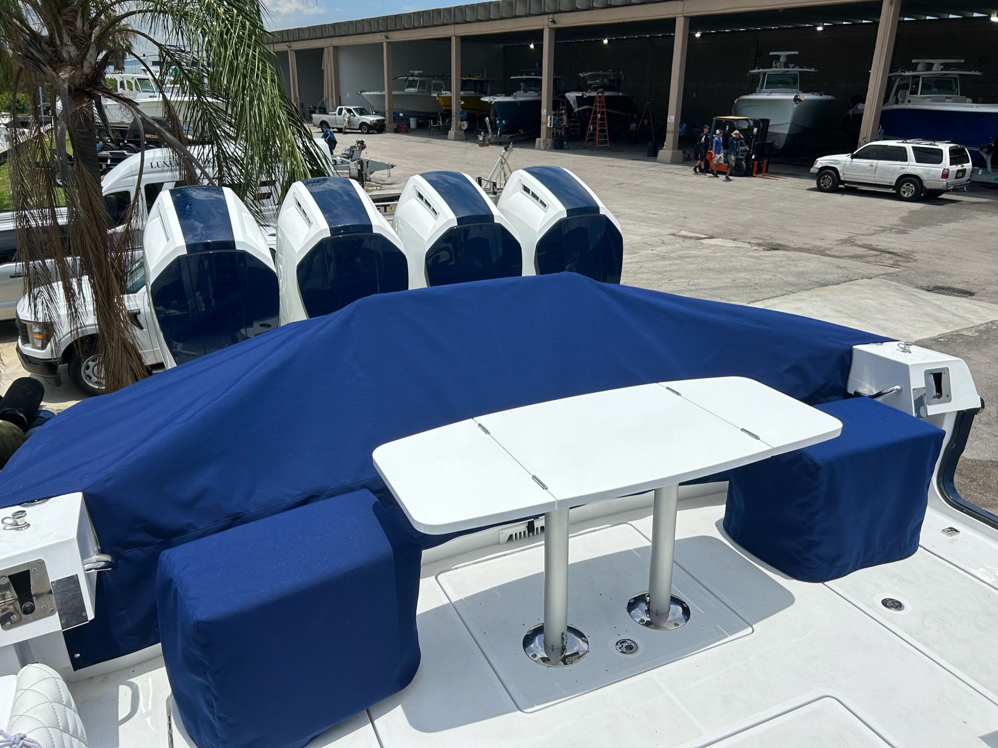Four white outboard motors and a boat stern fitted with navy blue canvas covers and a white folding table at a boatyard with covered boats in the background.