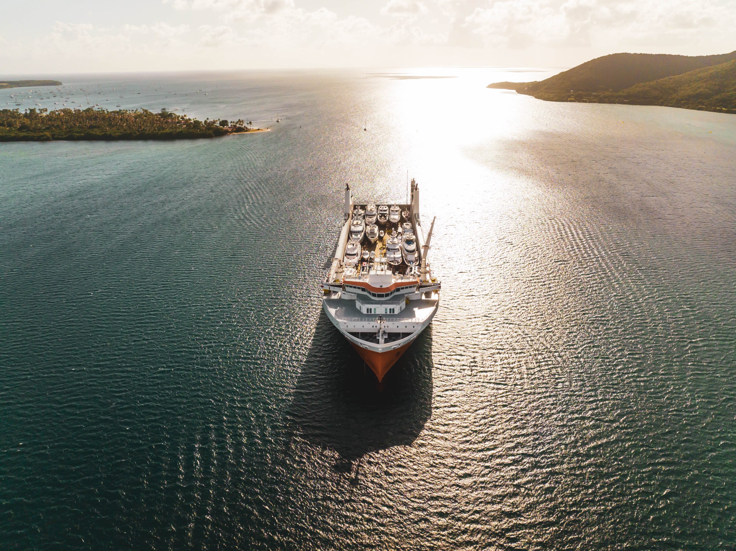 Aerial view of a yacht transport ship carrying several luxury yachts across a tropical bay at sunset.