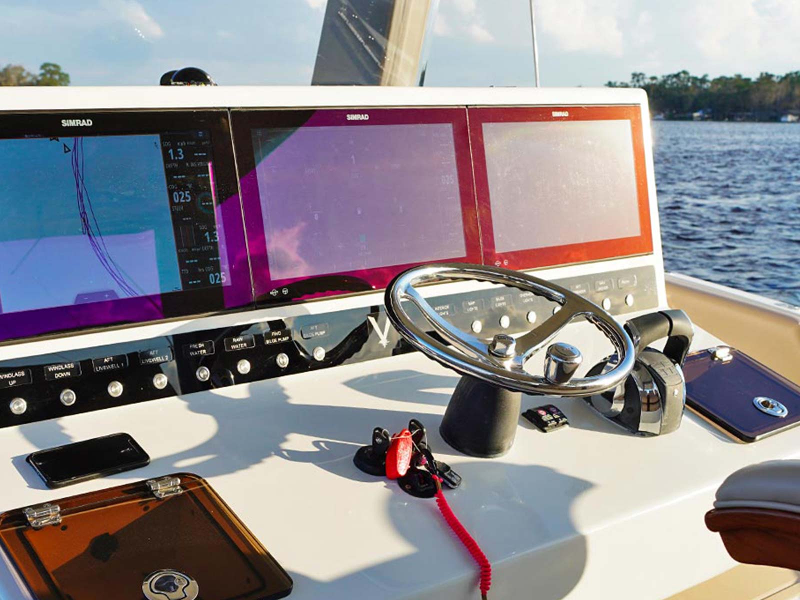 Boat helm with Simrad chartplotter displays, a stainless steel steering wheel and a switch panel overlooking a waterway.