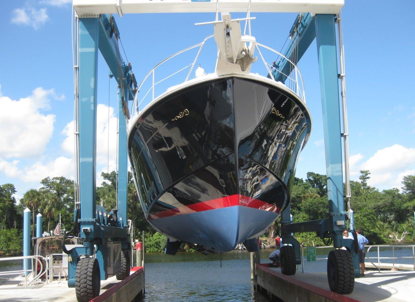 Black-hulled yacht with a red boot stripe being lifted by a travel lift at a boatyard on a tree-lined waterway.