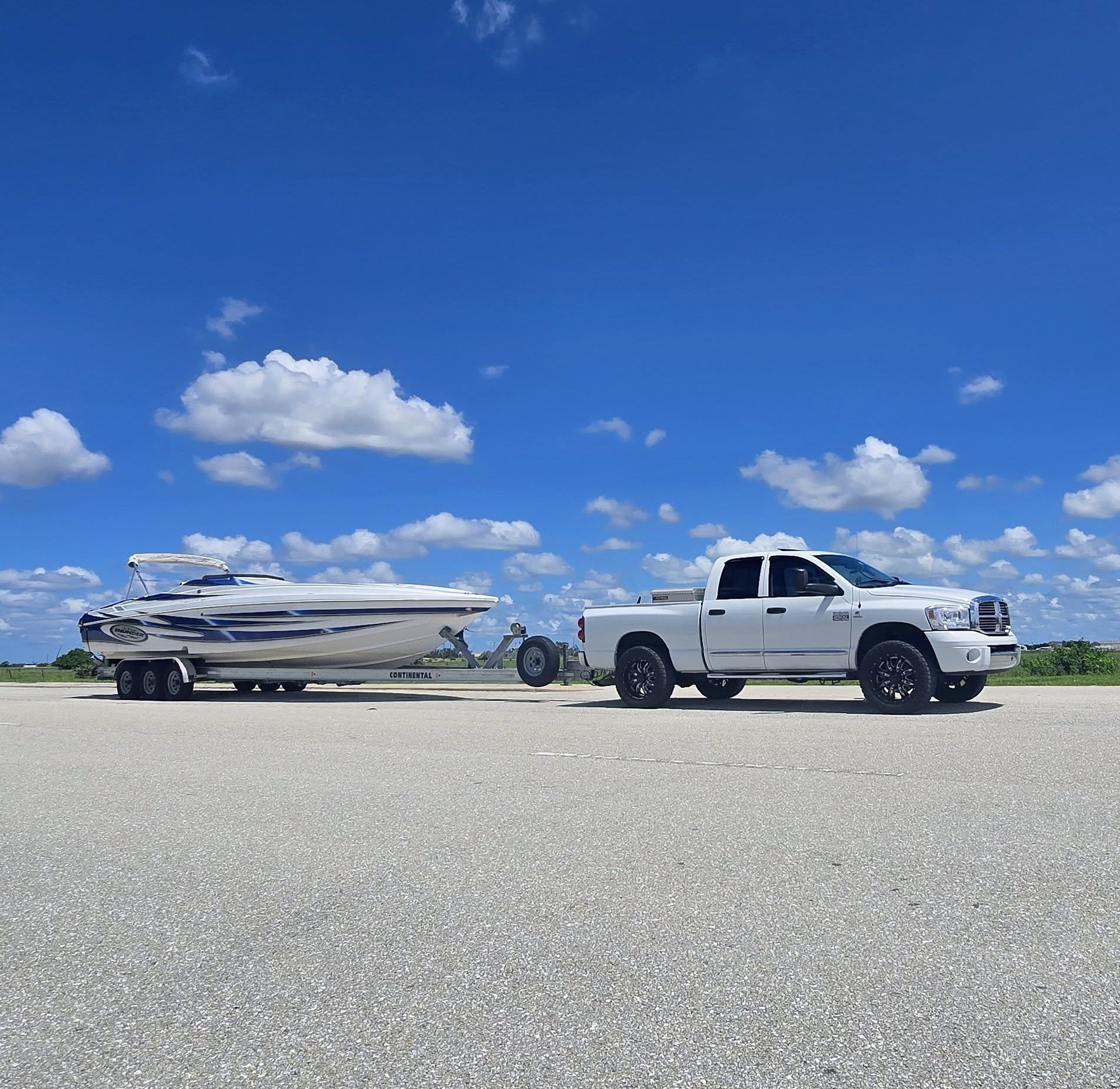 White speedboat with blue striping on a Continental trailer, towed by a white Dodge Ram pickup on a paved lot under a bright blue sky.