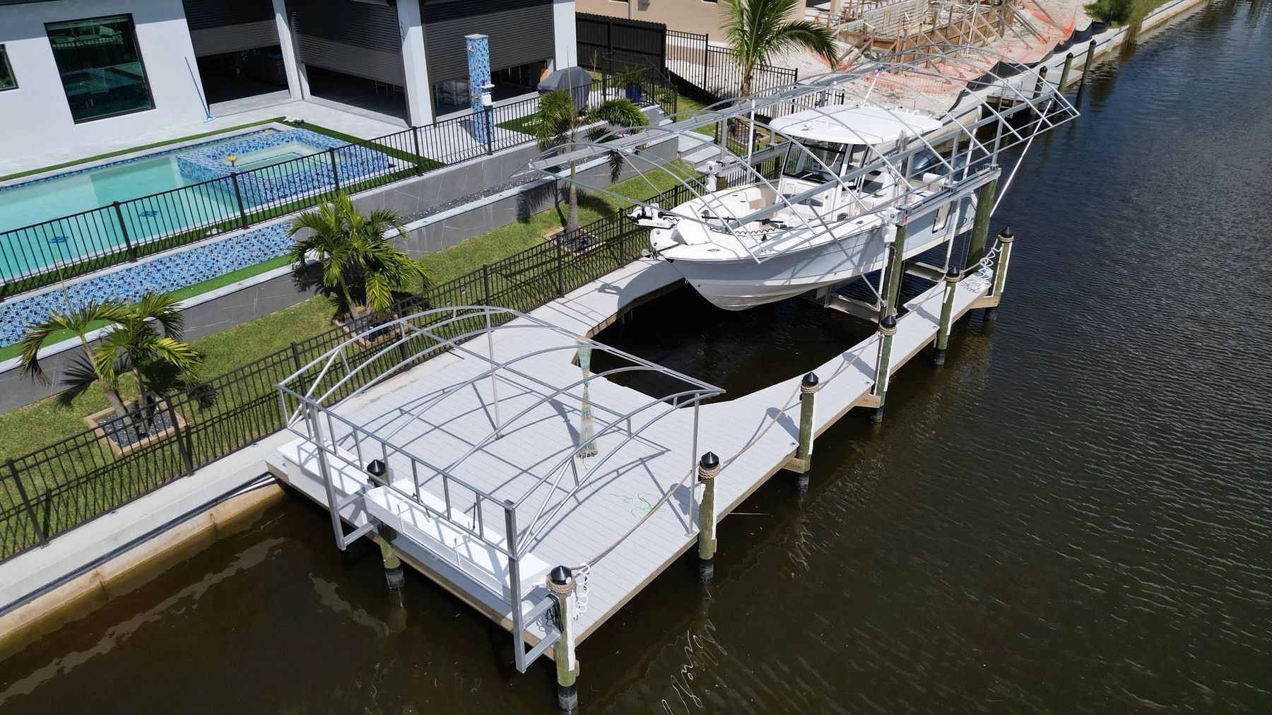 White sport fishing boat resting on a boat lift beside a grey composite dock on a residential canal, with a pool and palm trees on the bank.
