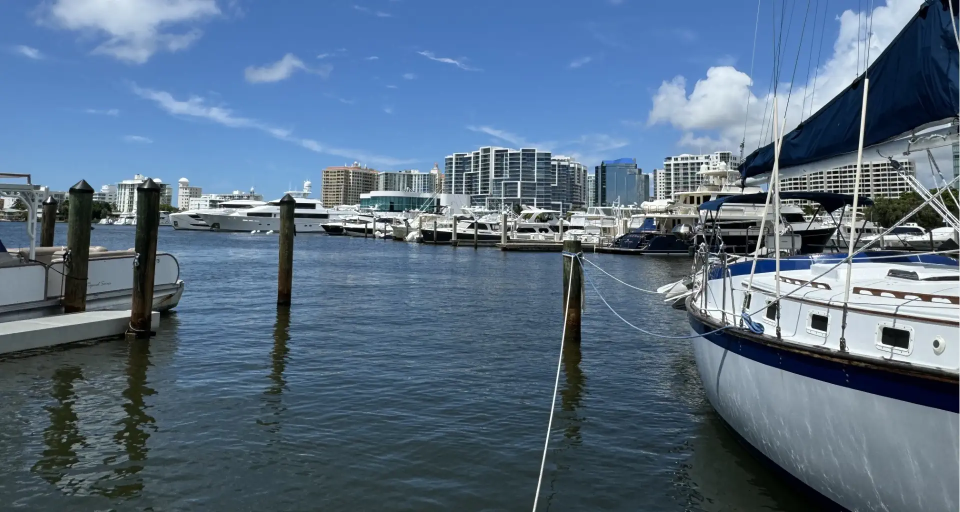 Sailboat and yachts docked in a marina basin with a high-rise waterfront skyline behind, location not confirmed.