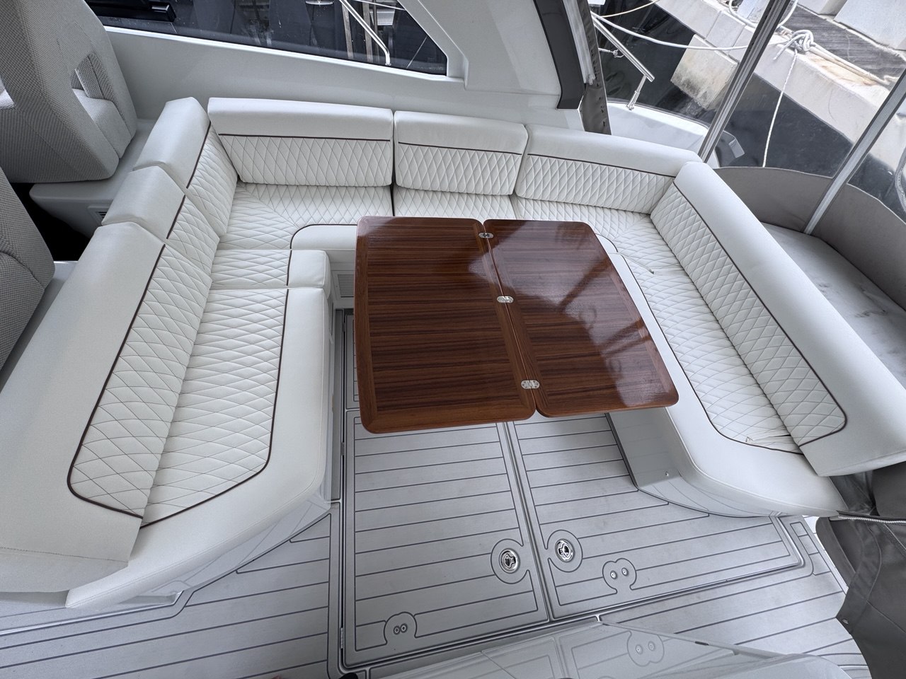 White diamond-quilted U-shaped lounge seating and a teak dining table inside a yacht cabin, with no specific business or town shown.