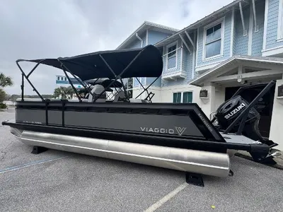 Black Viaggio pontoon boat with a Suzuki outboard motor and bimini top parked on asphalt in front of a blue-sided building.