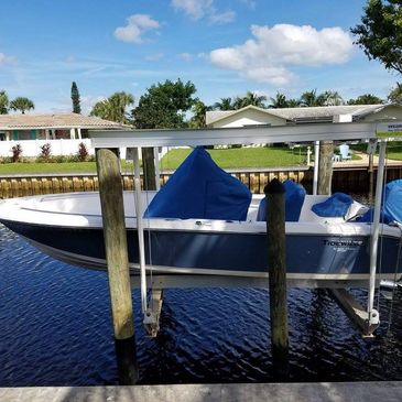Center console boat under a blue cover resting on a boat lift at a residential canal dock with palm trees and houses in the background.