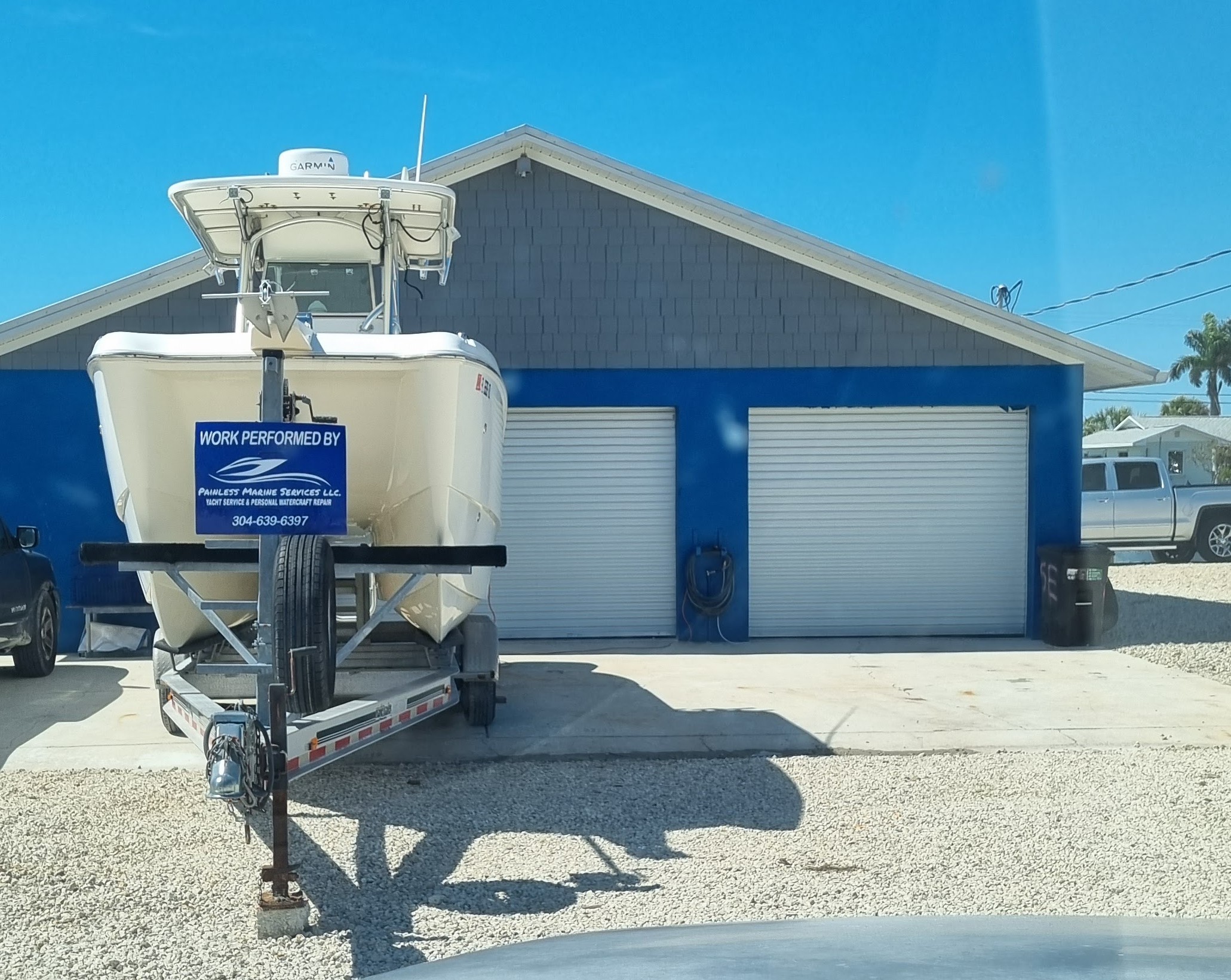 Center-console boat on a trailer displaying a 'Work Performed By Painless Marine Services LLC' sign in front of the company's blue garage building.