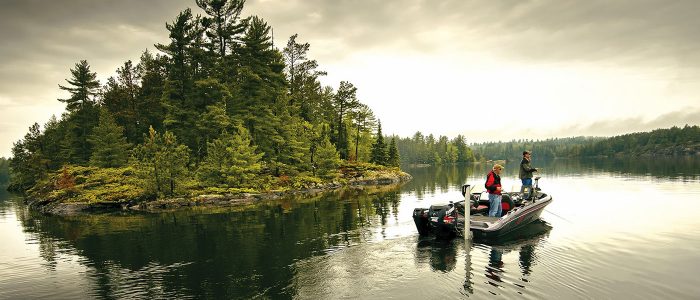 Two anglers fishing from a bass boat with twin outboard motors on a calm lake beside a pine-covered island.