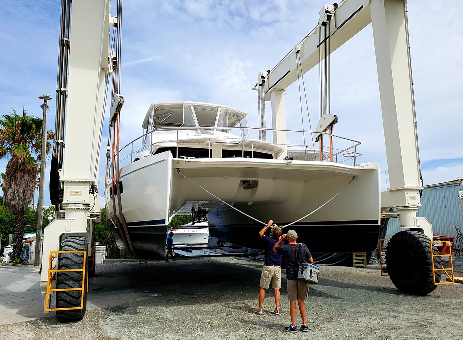 Catamaran being hoisted by a travel lift at a boatyard, with two people guiding the hull lines.