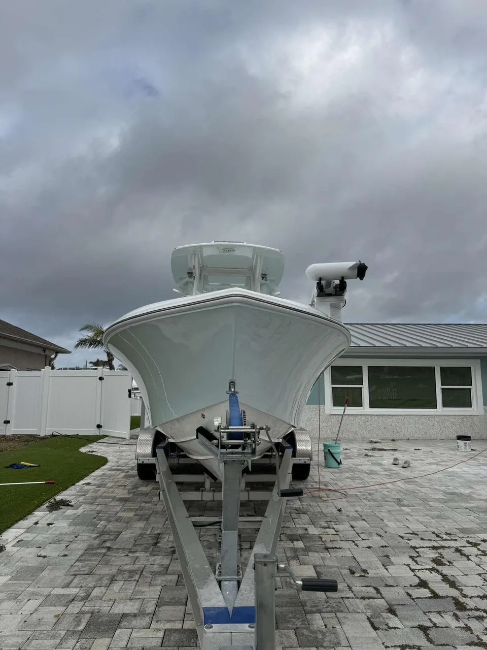 White center console boat sitting on a trailer in a paver driveway beside a house, with a bucket and hose nearby, suggesting a detailing or prep job in progress.