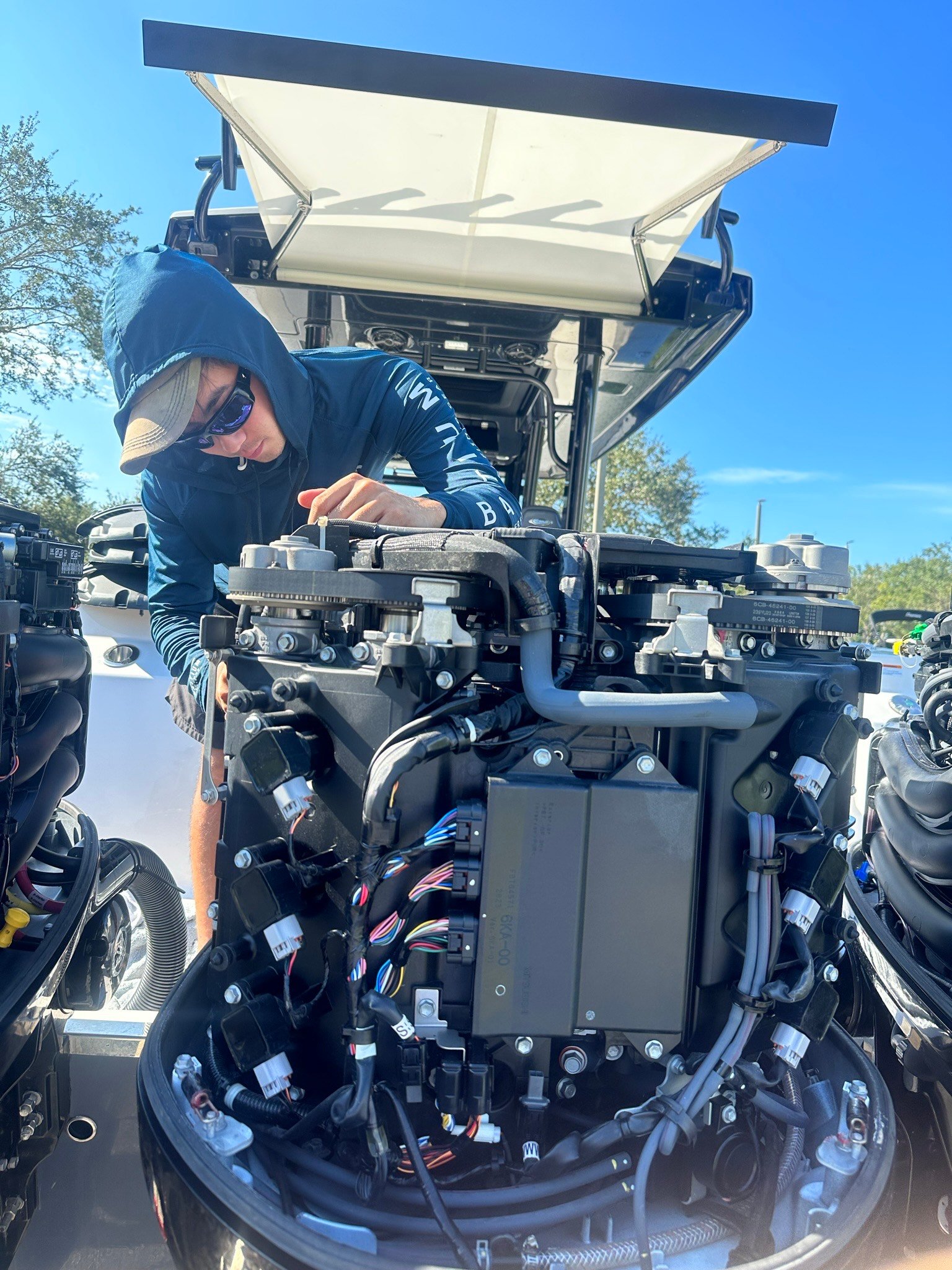Technician in a hooded blue shirt and cap working on the wiring and sensors of a multi-engine outboard motor mounted on a boat, outdoors under a clear sky.