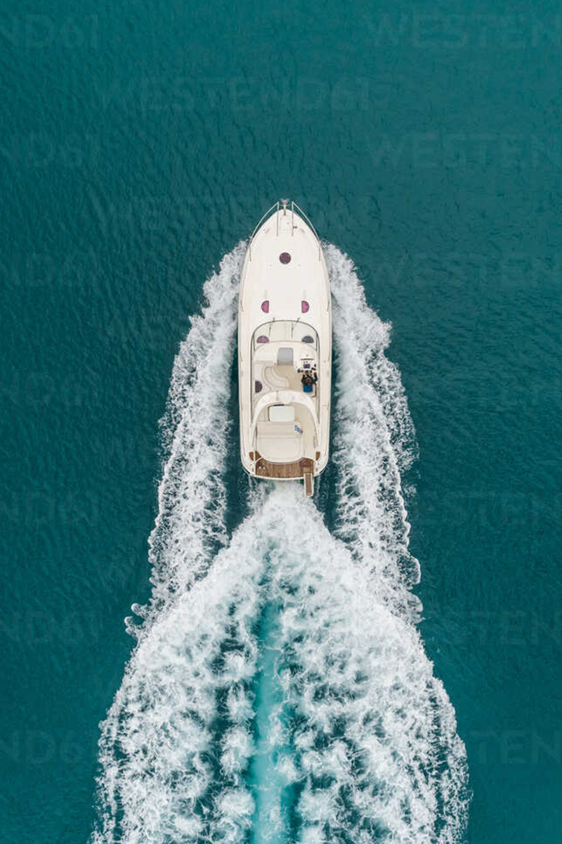 Aerial overhead photograph of a white motor yacht speeding across teal open water, leaving a foamy wake behind it.