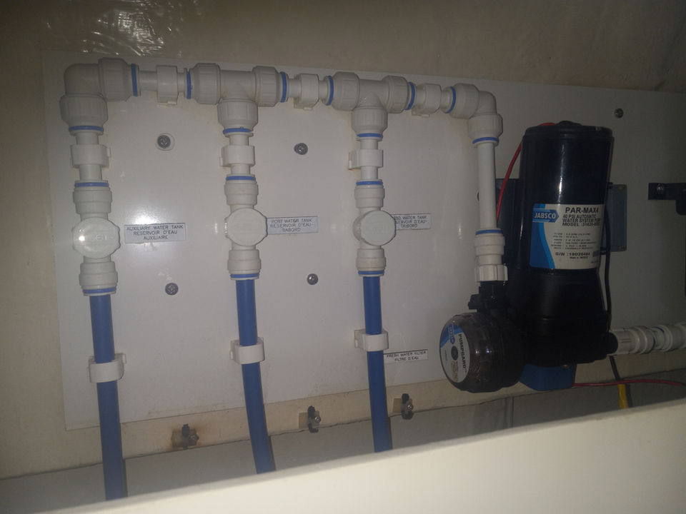 Freshwater tank manifold with blue supply lines and a Jabsco Par-Max pressure pump mounted on a white panel inside a boat.
