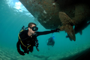 Scuba diver examining the underside of a boat hull in clear water over a sandy bottom.