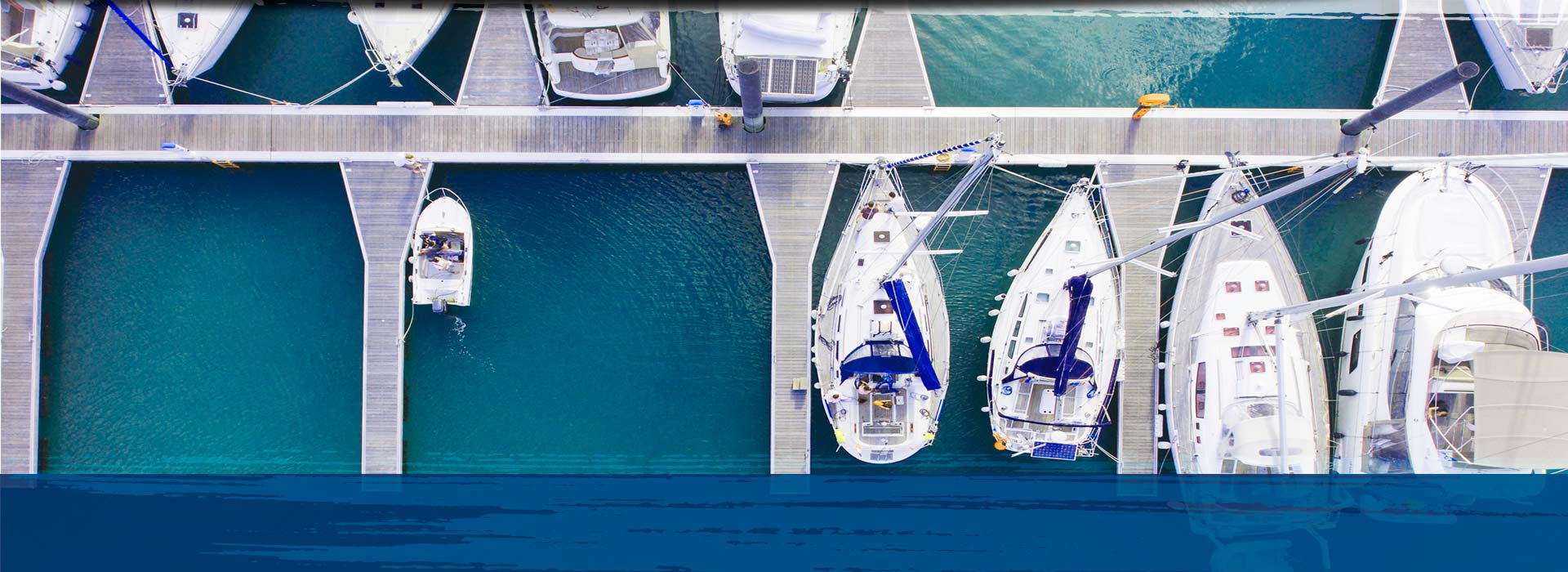 Aerial view of white sailboats and yachts moored at wooden docks in a marina with teal water.