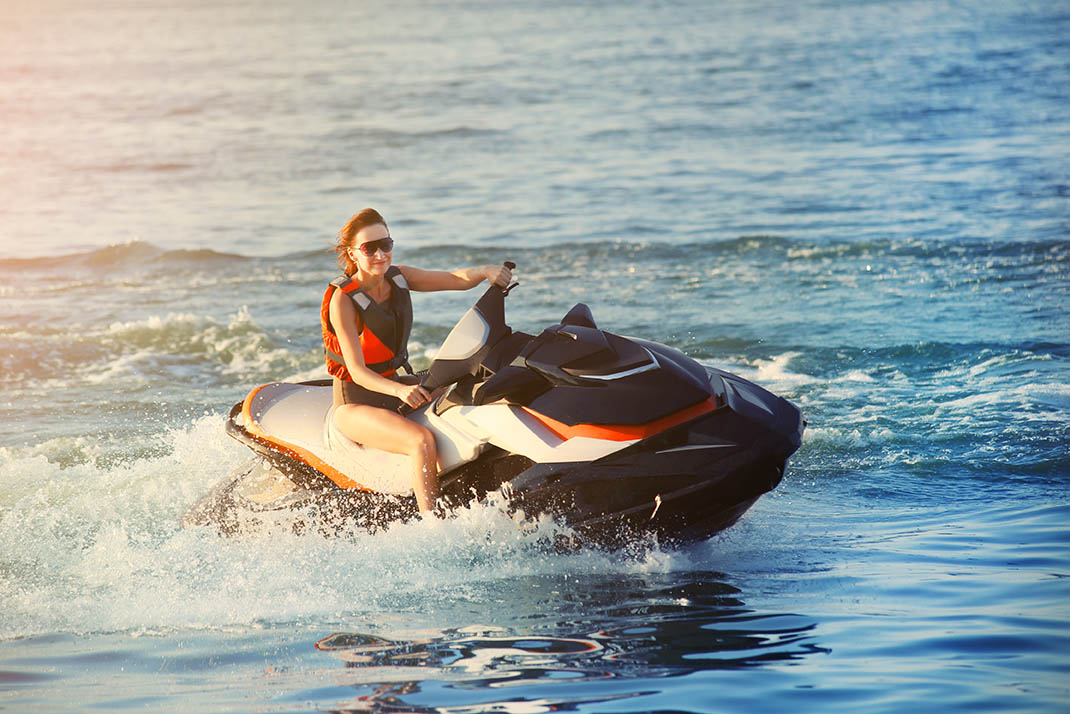 Woman in a life vest riding a white and orange personal watercraft across open water at sunset.