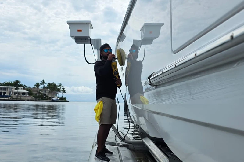Technician polishing the white hull of a yacht with a rotary buffer on a calm waterfront, with waterfront homes and palm trees in the background.