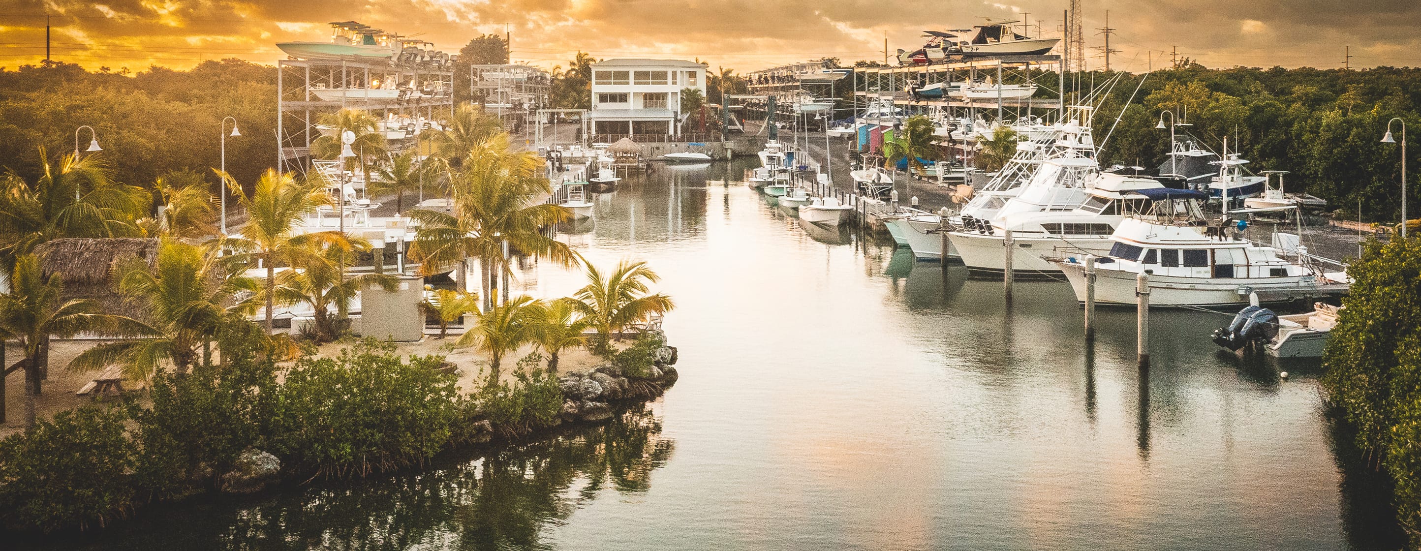 Sunset view of a canal lined with moored sportfishing boats, dry-stack boat storage racks, and palm trees at a marina in the Florida Keys.