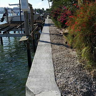 Concrete seawall walkway with boat docks and lifts along a canal, lined with flowering shrubs and gravel landscaping.