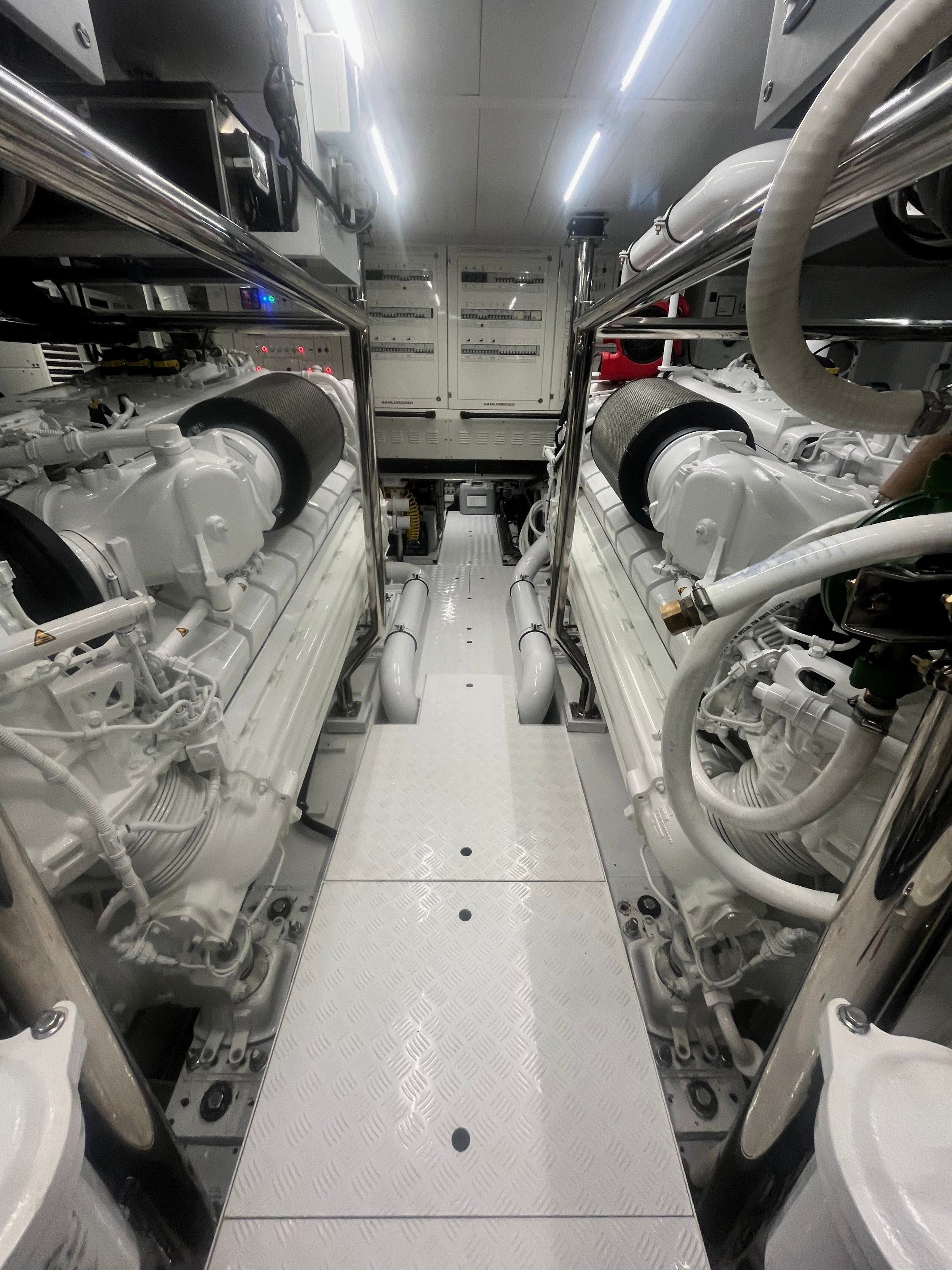 Twin white diesel main engines with polished stainless railings and a SanLorenzo generator switchboard in a yacht engine room, location not identified.