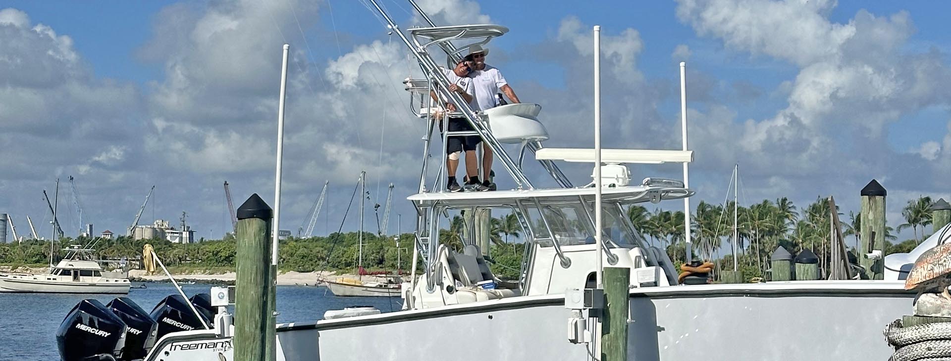 Two crew members on the T-top of a white Freeman sportfishing boat with Mercury outboard engines docked at a marina.