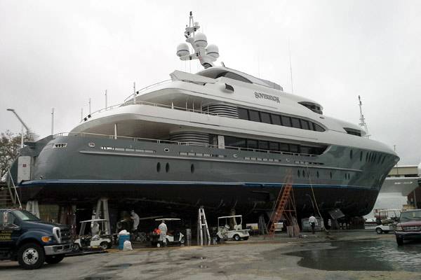 Large grey motor yacht Sentinel on stands during refit at a boatyard, with workers and yard vehicles nearby.