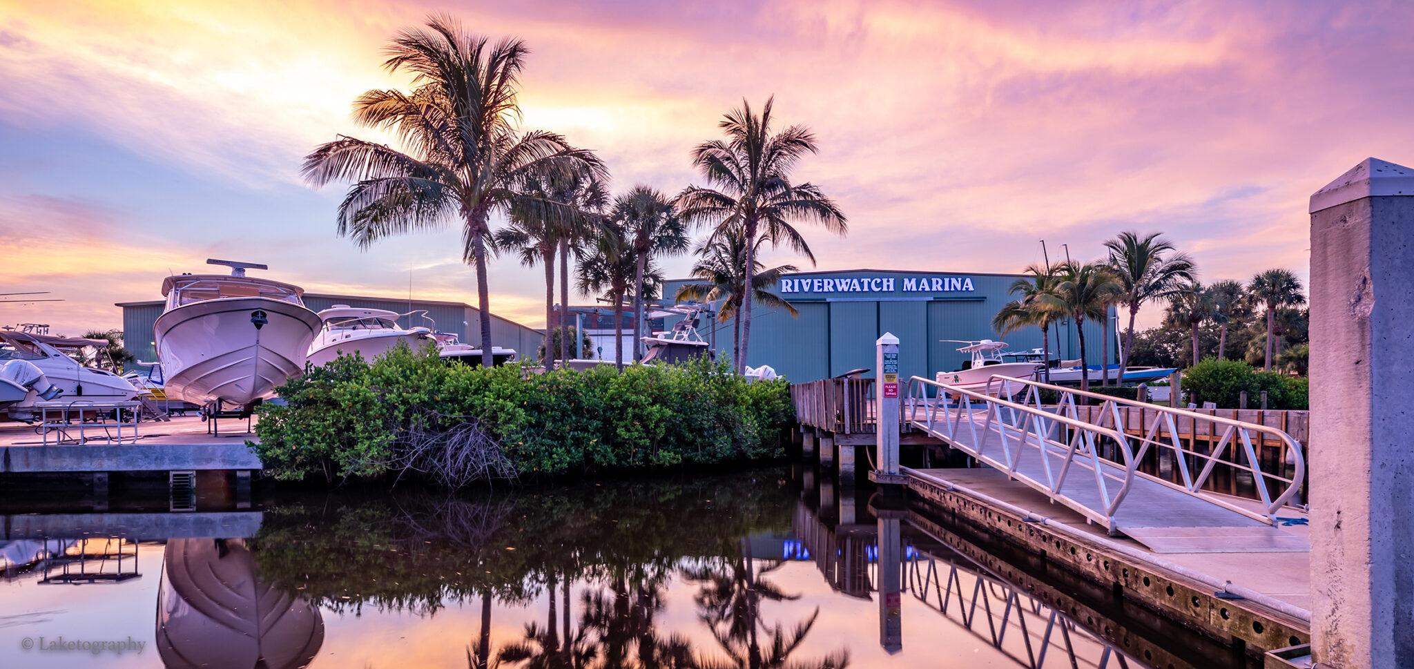 Boats in dry storage and a floating dock with a gangway at Riverwatch Marina, a waterfront facility lined with palm trees at sunset.