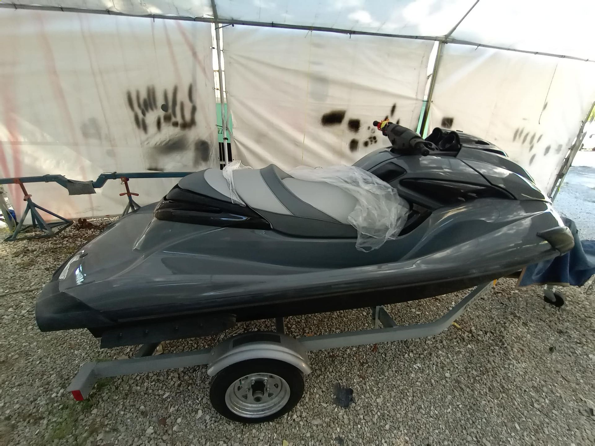 A gray personal watercraft with a light gray seat, wrapped in protective plastic, sits on a trailer under a white canopy on gravel.