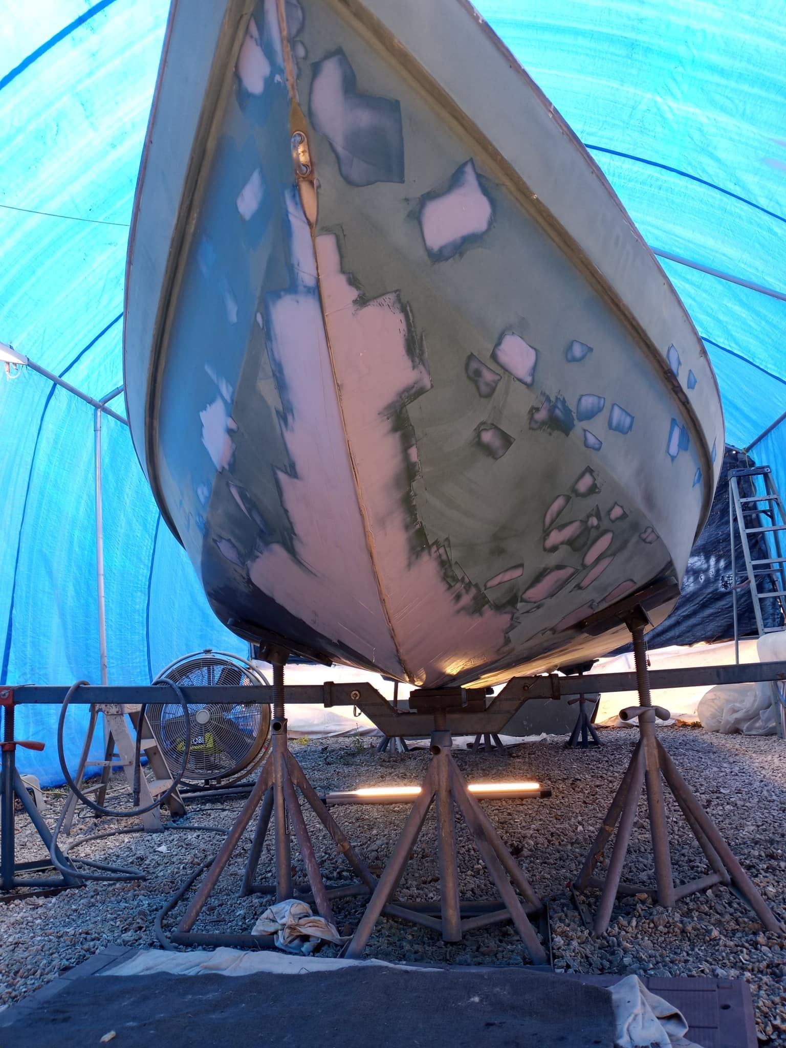Fiberglass boat hull on jack stands under a blue tarp enclosure, with repair patches being worked on.
