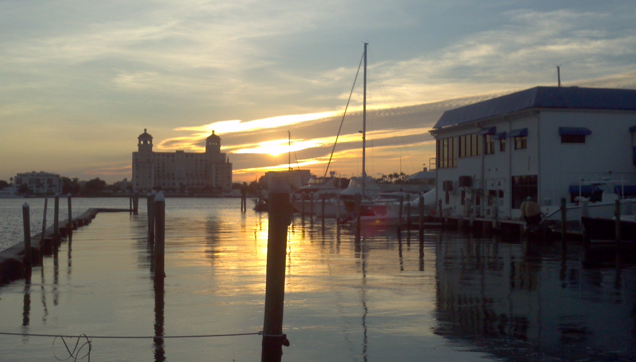 Sunset over a marina with docked sailboats, powerboats, and pilings beside a white waterfront building, with a tower hotel in the distance.