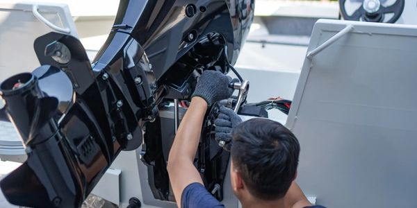 Technician servicing an outboard motor on a boat, with the engine's lower unit and wiring visible.