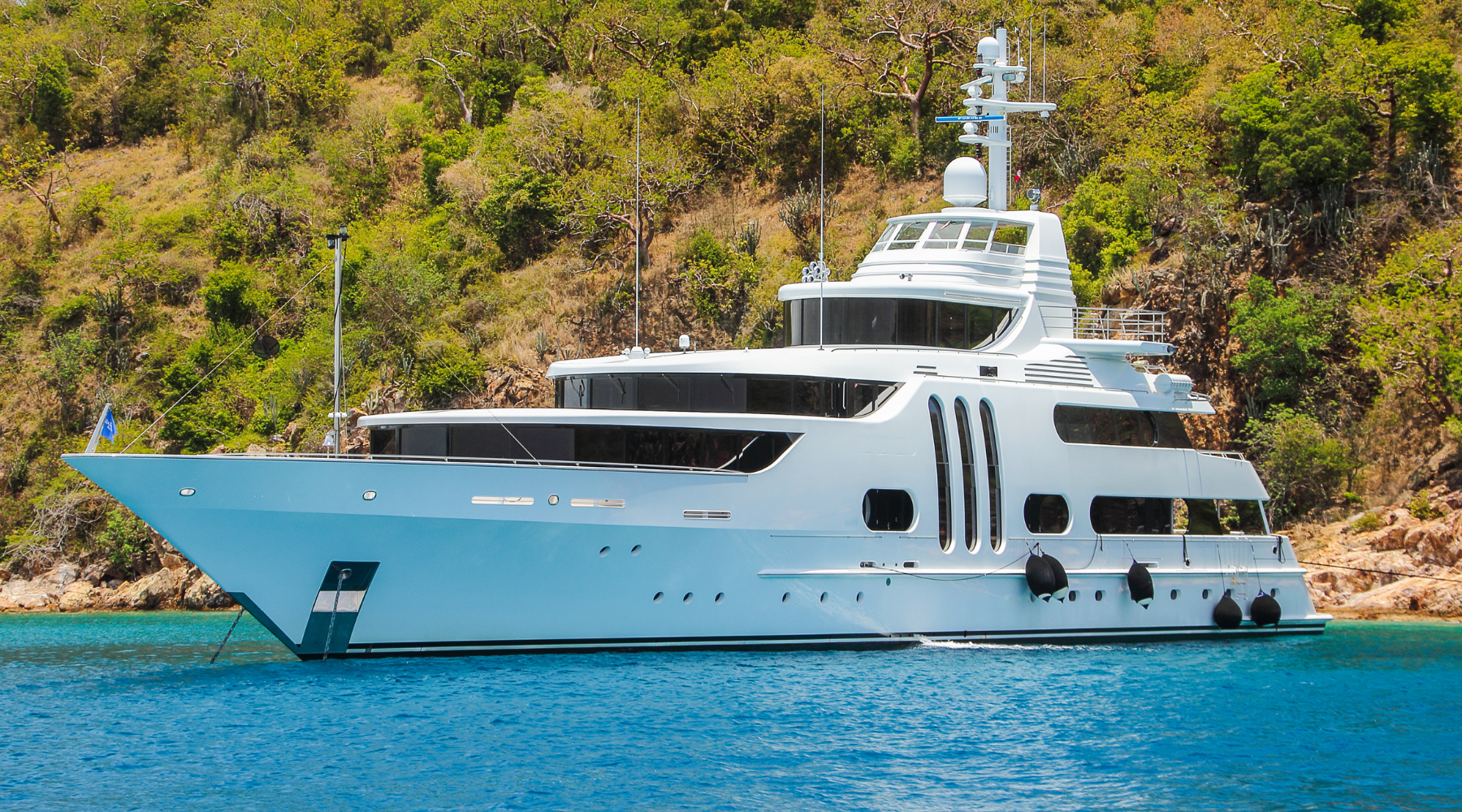 Large white motor yacht at anchor in a turquoise bay below a green hillside, with its name and town not visible in the photo.