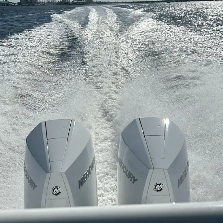 Twin Mercury outboard motors on a boat leaving a white wake across open water with a shoreline of buildings in the distance.