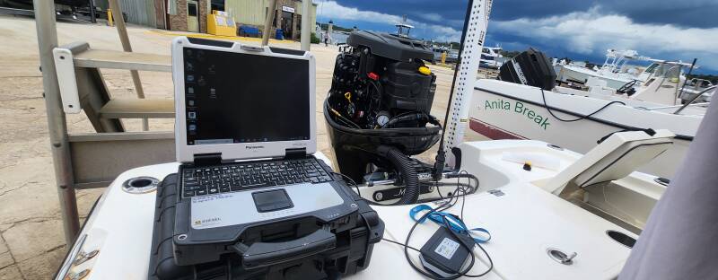 Rugged laptop connected to an outboard motor for diagnostics on a boat deck at a marina dock, with the boat Anita Break alongside.