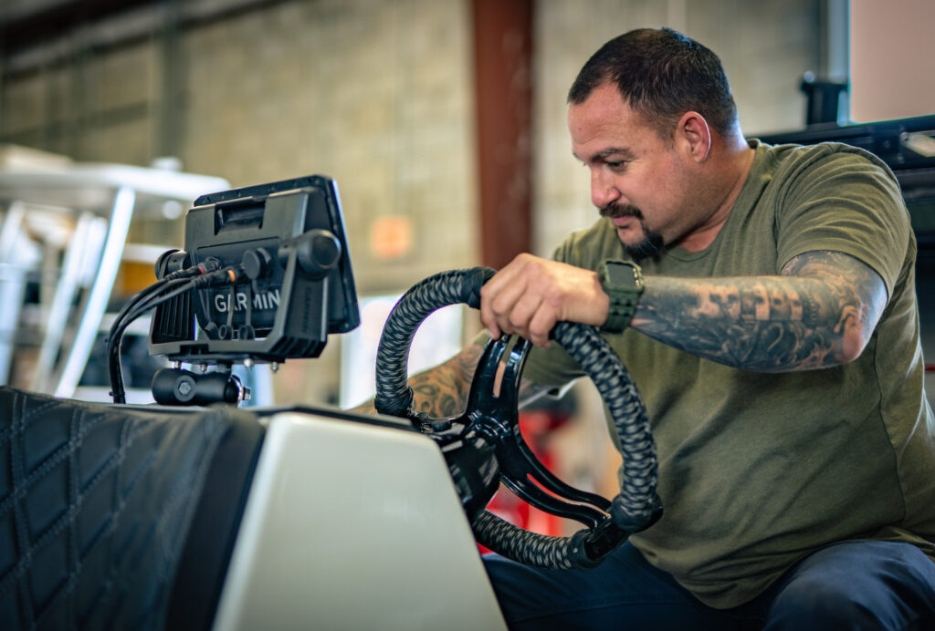 Technician adjusting a Garmin marine chartplotter and rope-wrapped steering wheel at a boat helm in a workshop.
