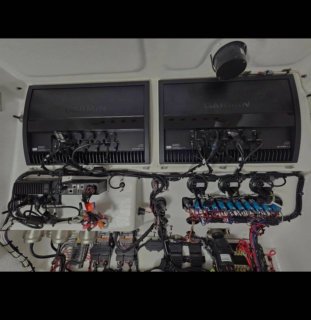 Two Garmin marine chartplotter displays with a dense wiring harness, breakers, and electrical panels mounted inside a boat's electronics compartment.