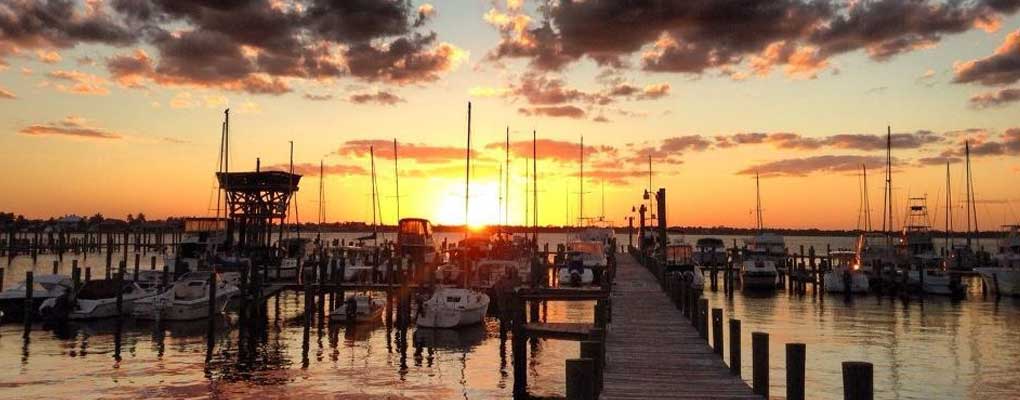 Sailboats and powerboats moored at a marina dock during sunset, with a wooden walkway extending into the calm water.
