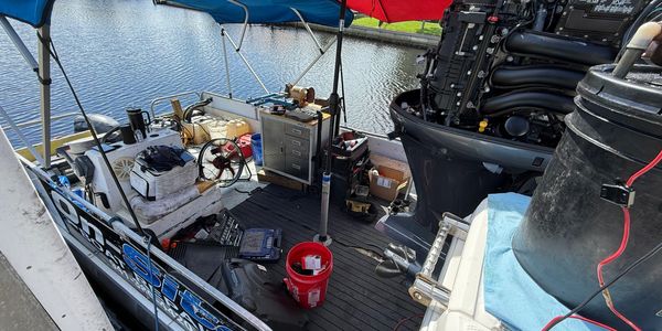Outboard motor and workdeck of a boat docked on calm water, with tools, a red bucket, and wiring laid out during maintenance.