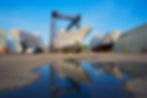 Shipyard hulls and a gantry crane reflected in a puddle on a quay.