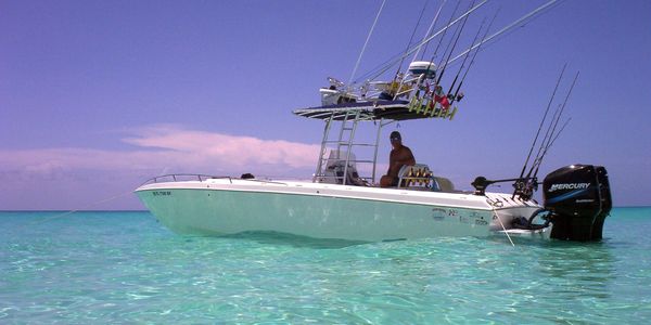 Center console fishing boat with a Mercury outboard and rigged rod holders anchored in shallow turquoise water, with a crew member aboard.