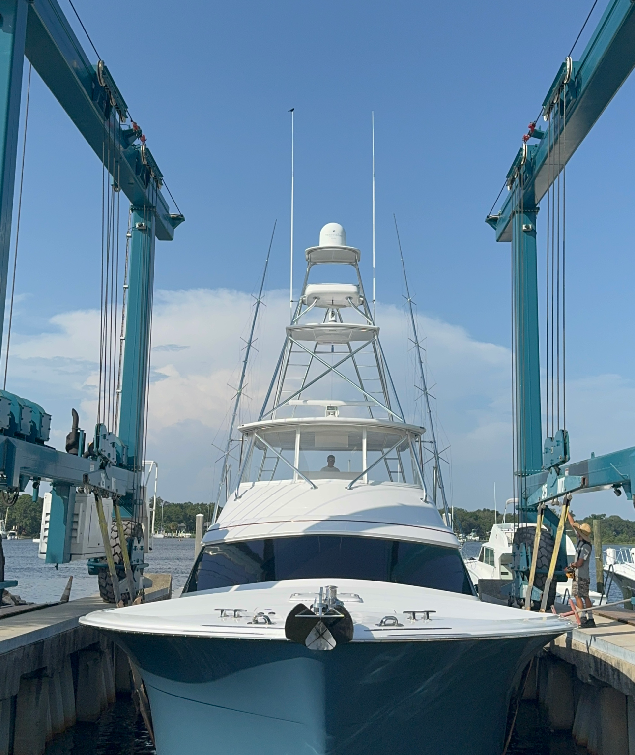 Large white sportfishing boat with a tower and radar being lifted by a teal travel lift at a boatyard on the water.