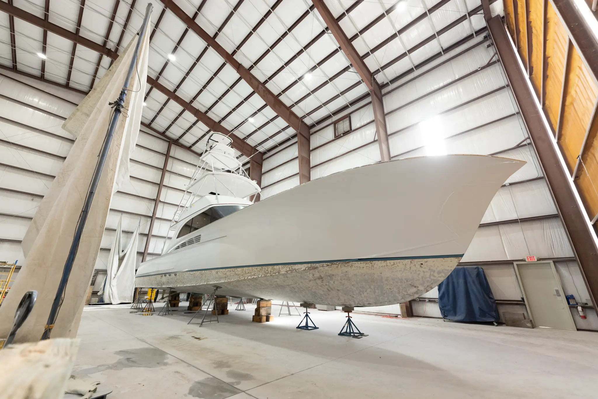 Large white sport-fishing boat on jack stands inside an indoor boatyard shed, with its hull showing wear and bottom work underway.